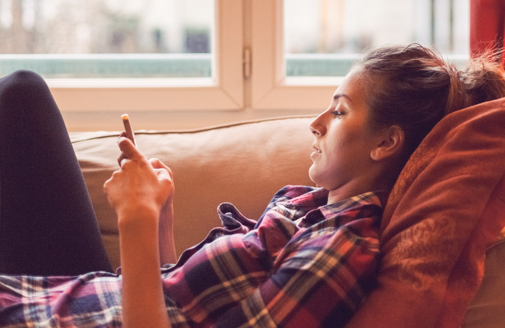 Kvinna som ligger i en soffa hemma och kollar i en mobil